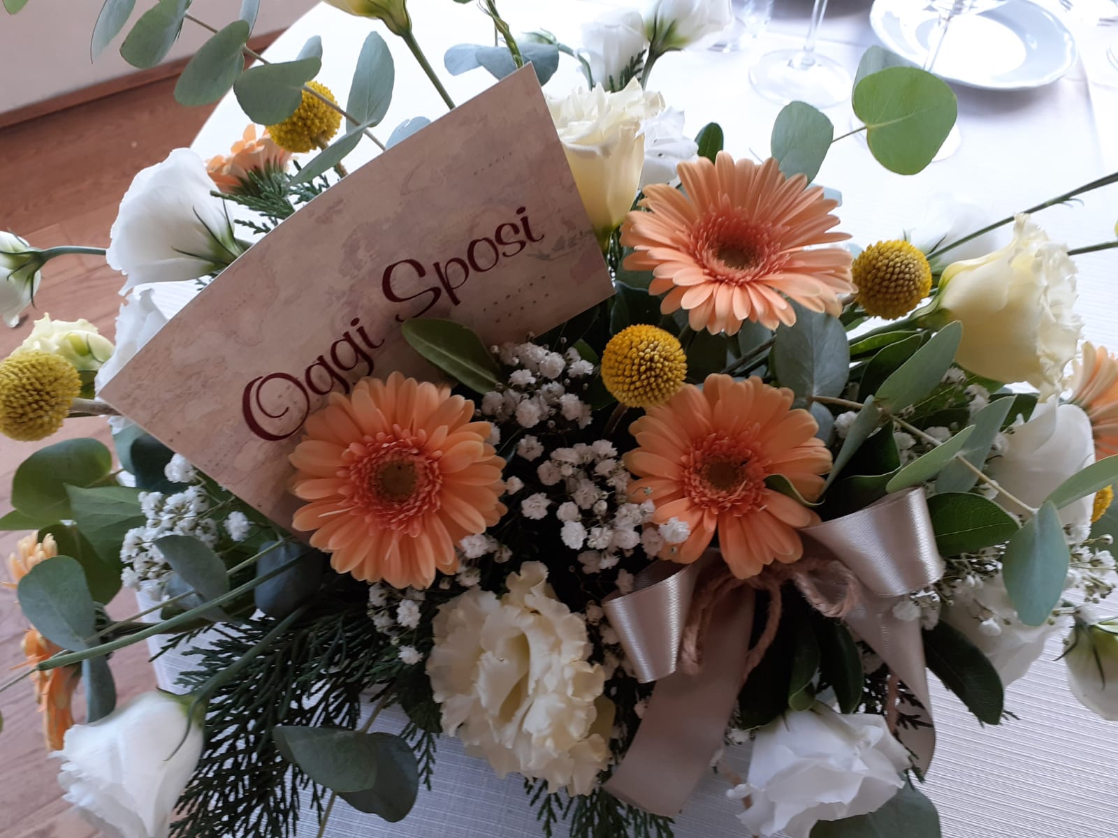 Composizione floreale con margherite Gerber arancioni, fiori bianchi e un biglietto con la scritta