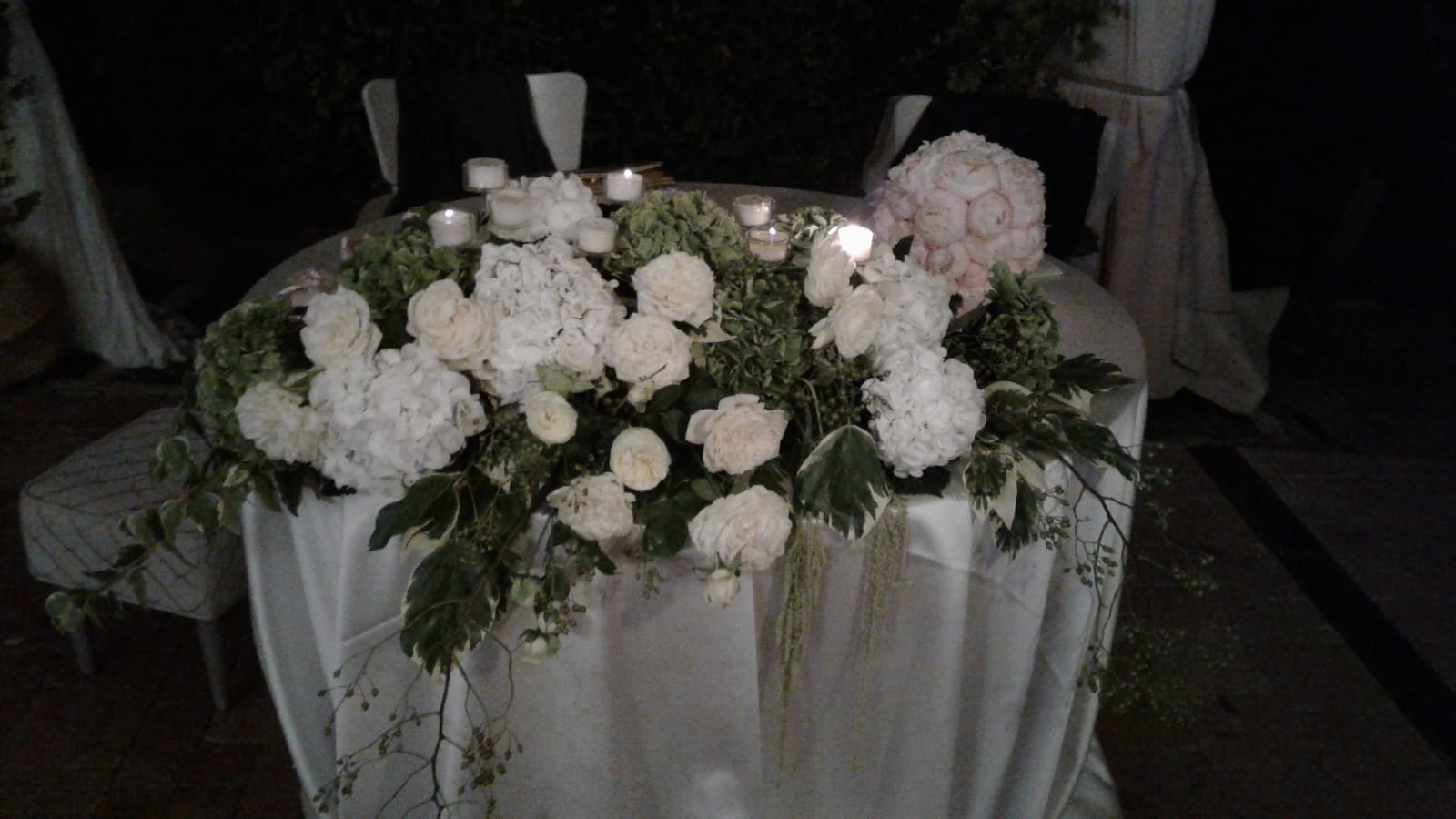 Tavolo apparecchiato per un evento con composizione floreale bianca e candele.