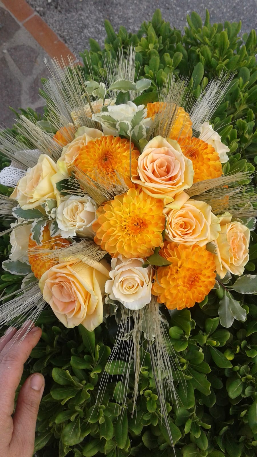 Bouquet di fiori gialli e color pesca con rametti ornamentali simili a spighe di grano, disposti a mano su fogliame verde.