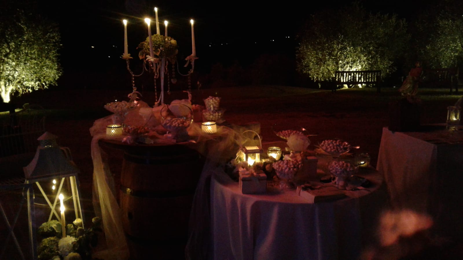 Tavolo da dessert illuminato da candele all'aperto di notte, con un grande candelabro e piccole lanterne.