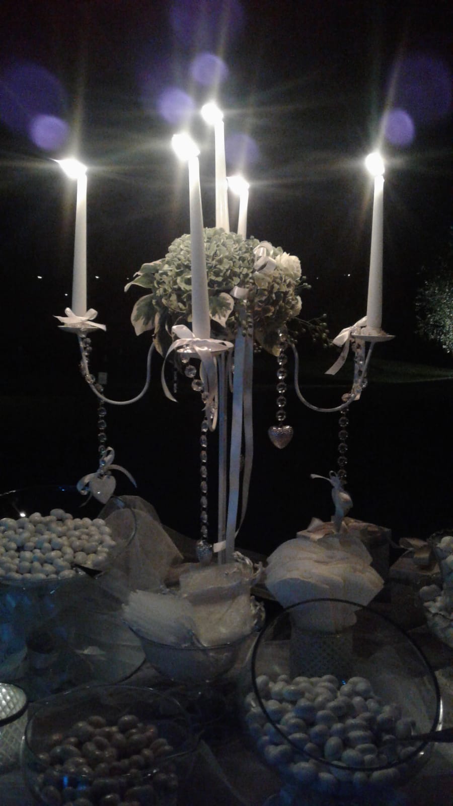 Centrotavola portacandele con candele bianche, fiori e cristalli su un tavolo con dolciumi.