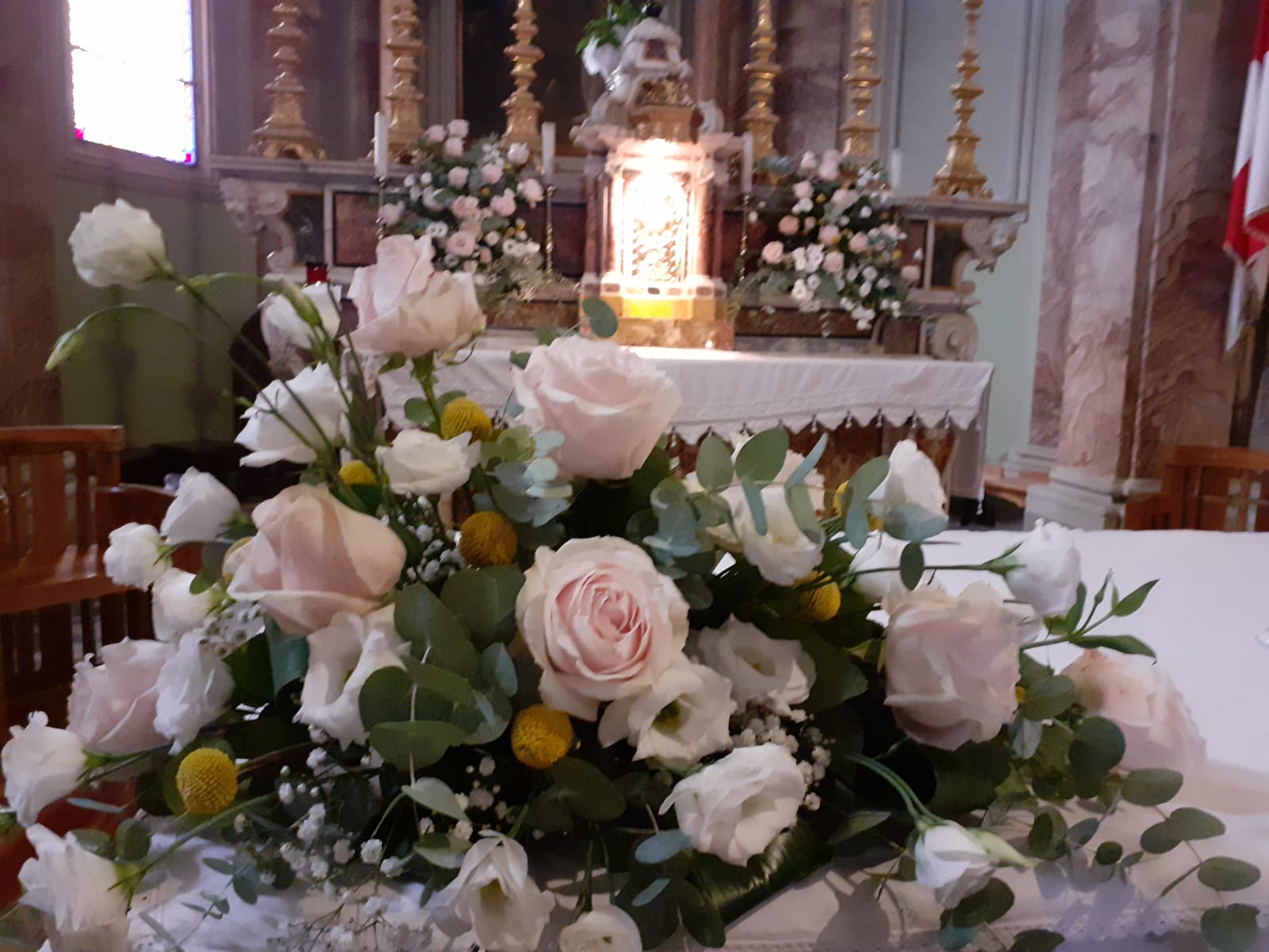 Composizione floreale di rose rosa e bianche, eucalipto e accenti gialli su un altare bianco in una chiesa.