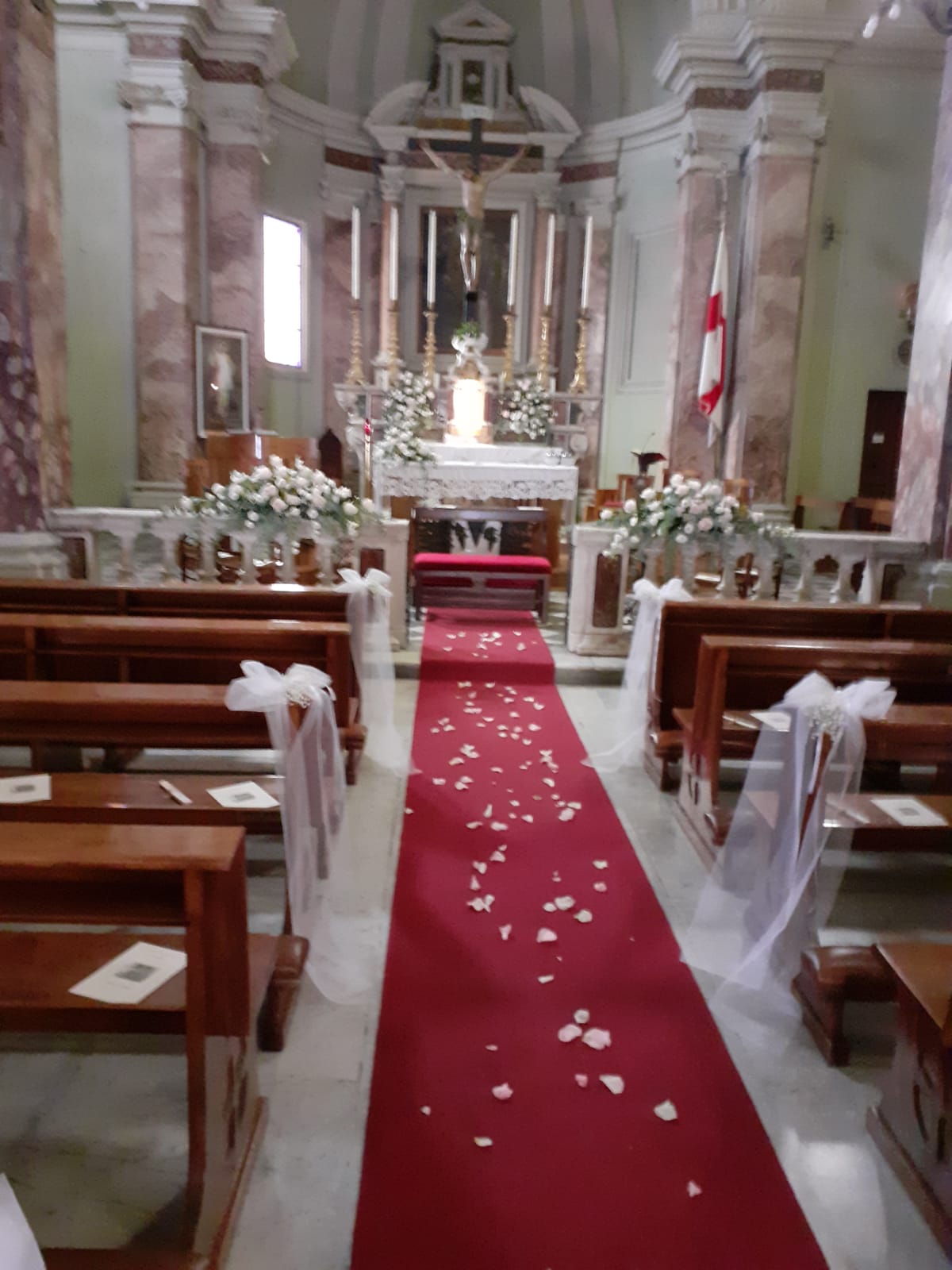 Vista interna di una chiesa decorata per un matrimonio, con navata centrale ricoperta di tappeto rosso e composizioni floreali.
