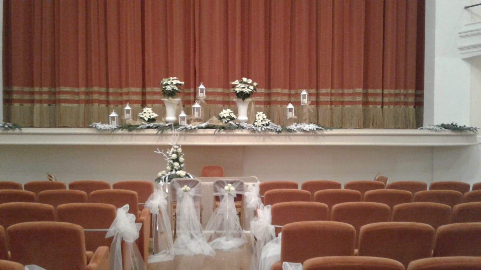 Auditorium decorato per una cerimonia nuziale: fiori, candele e tessuti sul palco e sulle sedie.