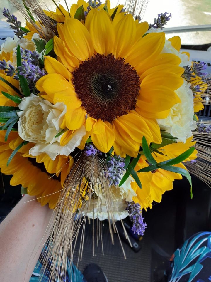 Mano che regge un mazzo di girasoli giallo brillante, fiori bianchi e dettagli di lavanda.