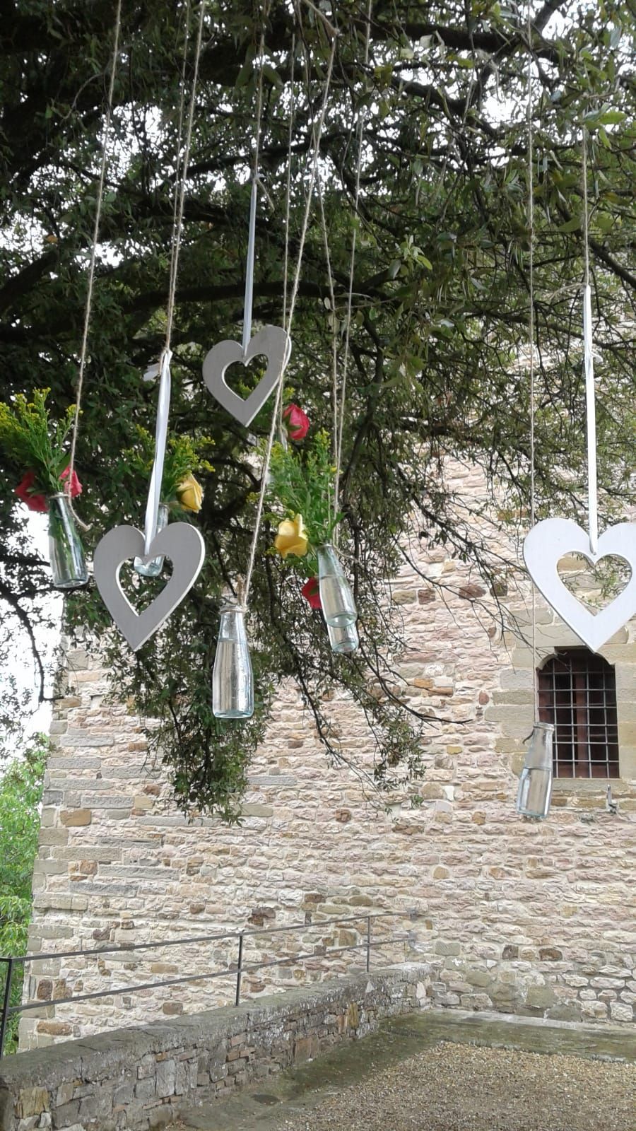 Decorazioni appese a un albero: cuori bianchi, bottiglie di vetro con fiori, contro un edificio in pietra.