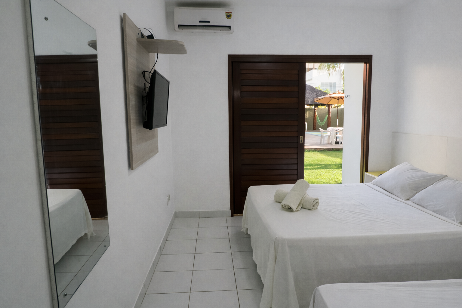 Quarto de hotel branco com cama, TV, espelho e porta que dá para um pátio.