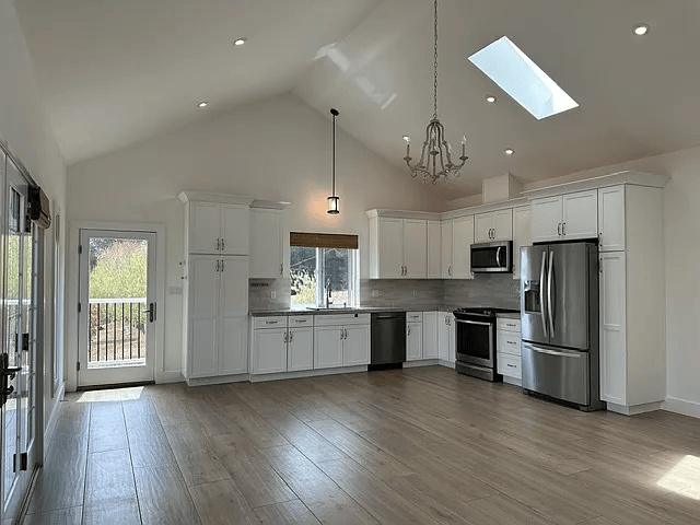 Bright, empty open-plan kitchen and living room with white cabinets, stainless appliances, and vaulted ceilings.