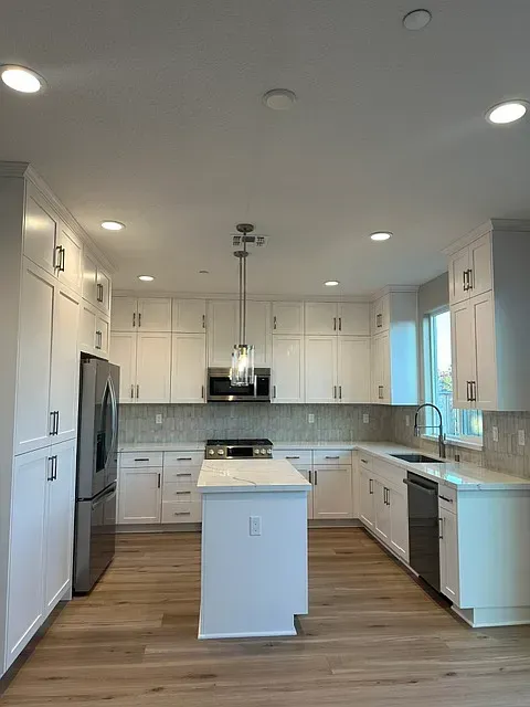 Bright white modern kitchen with island, stainless appliances, wood floors, and recessed lighting.