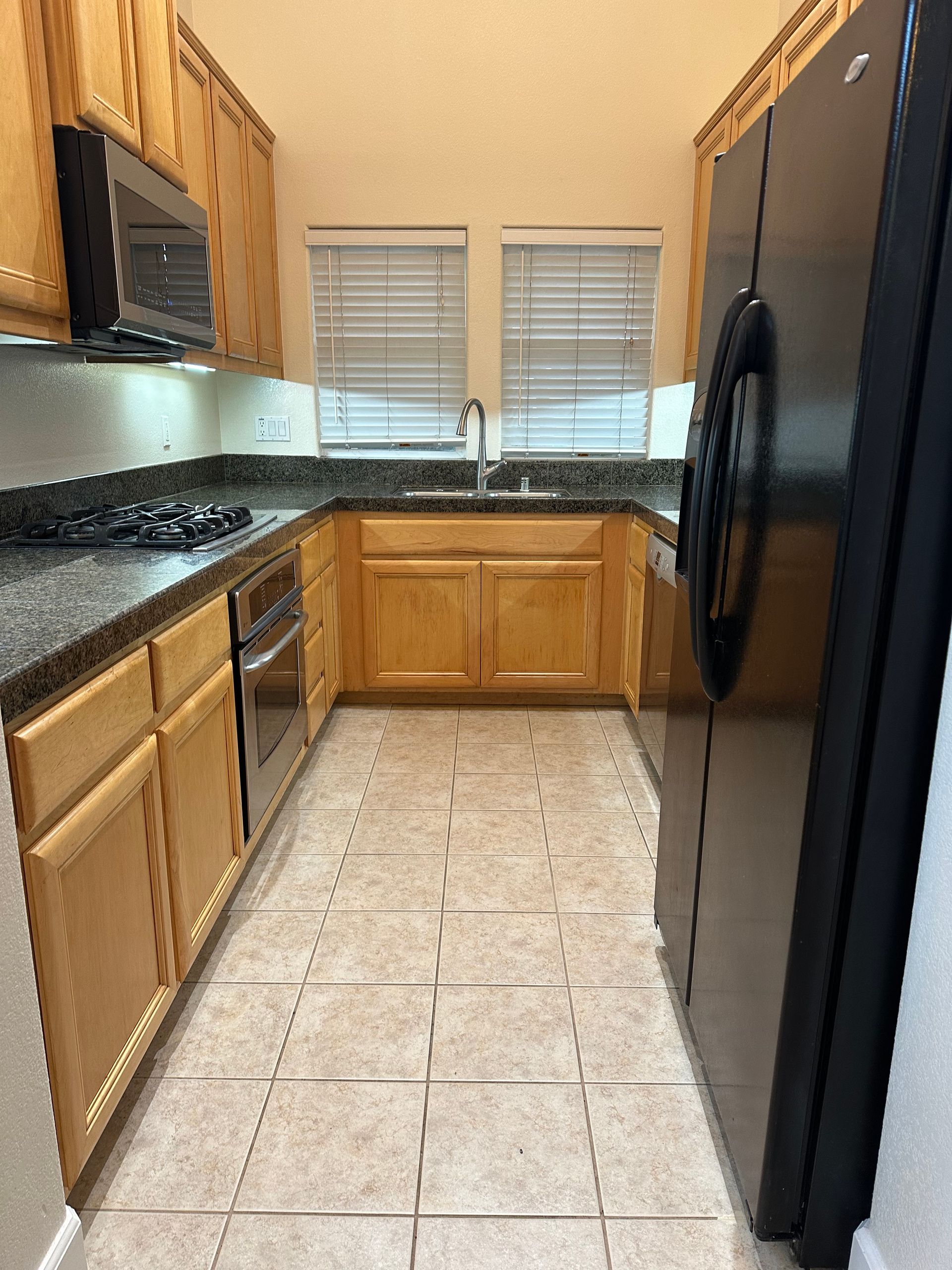 Narrow kitchen with light wood cabinets, black appliances, and tile floor, viewed from the doorway