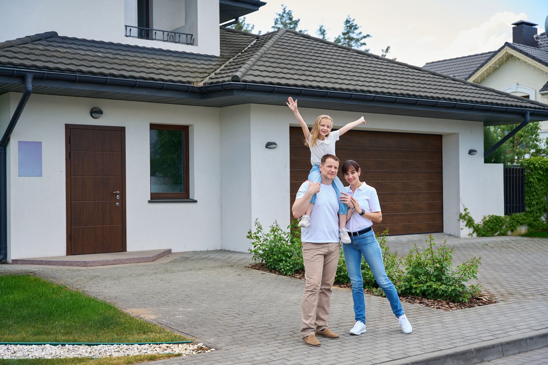A family stands on a paved driveway in front of their suburban home, with a child sitting on a parent's shoulders.