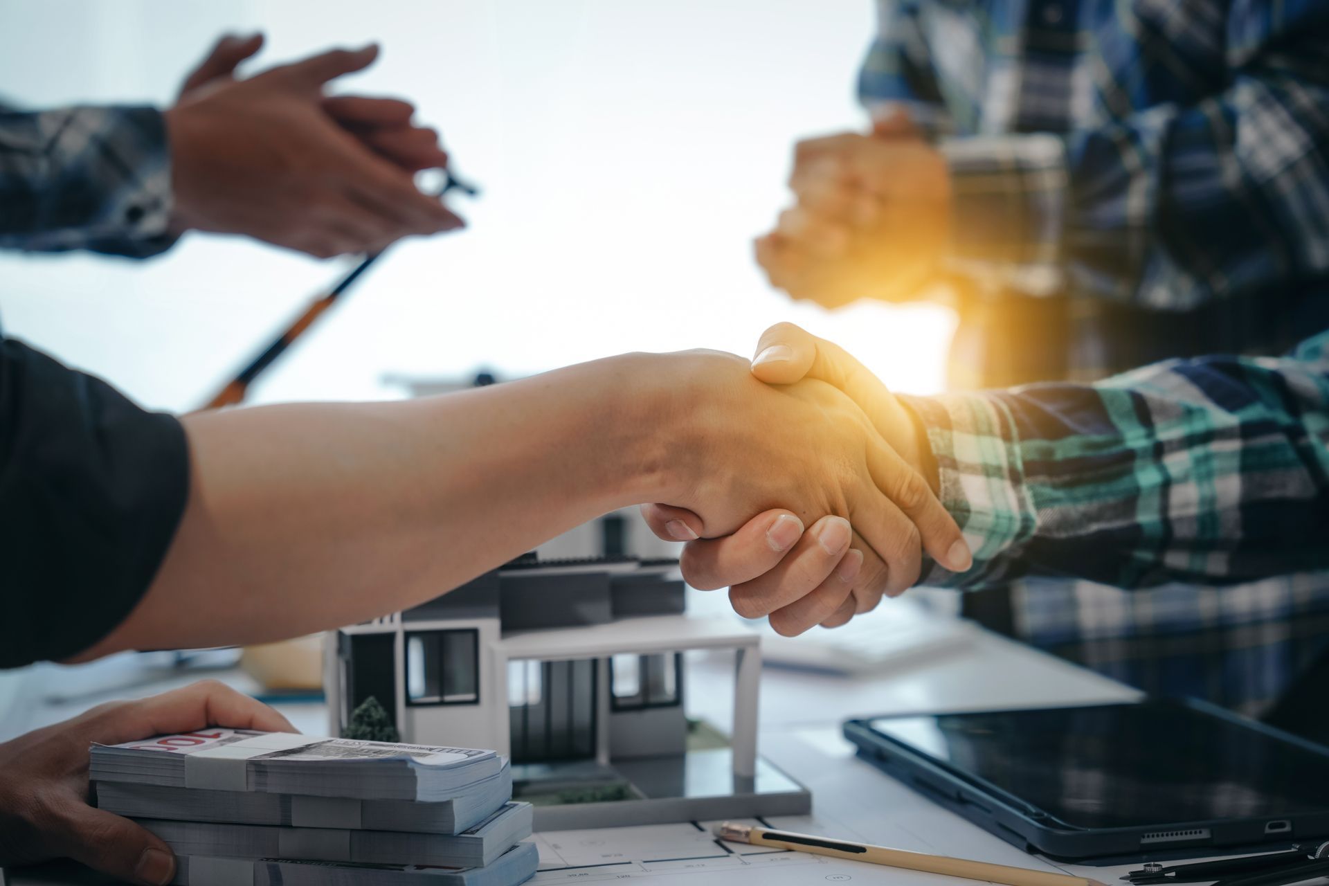 Two people shake hands over a table with a house model, a stack of cash, and a tablet, symbolizing a real estate deal.