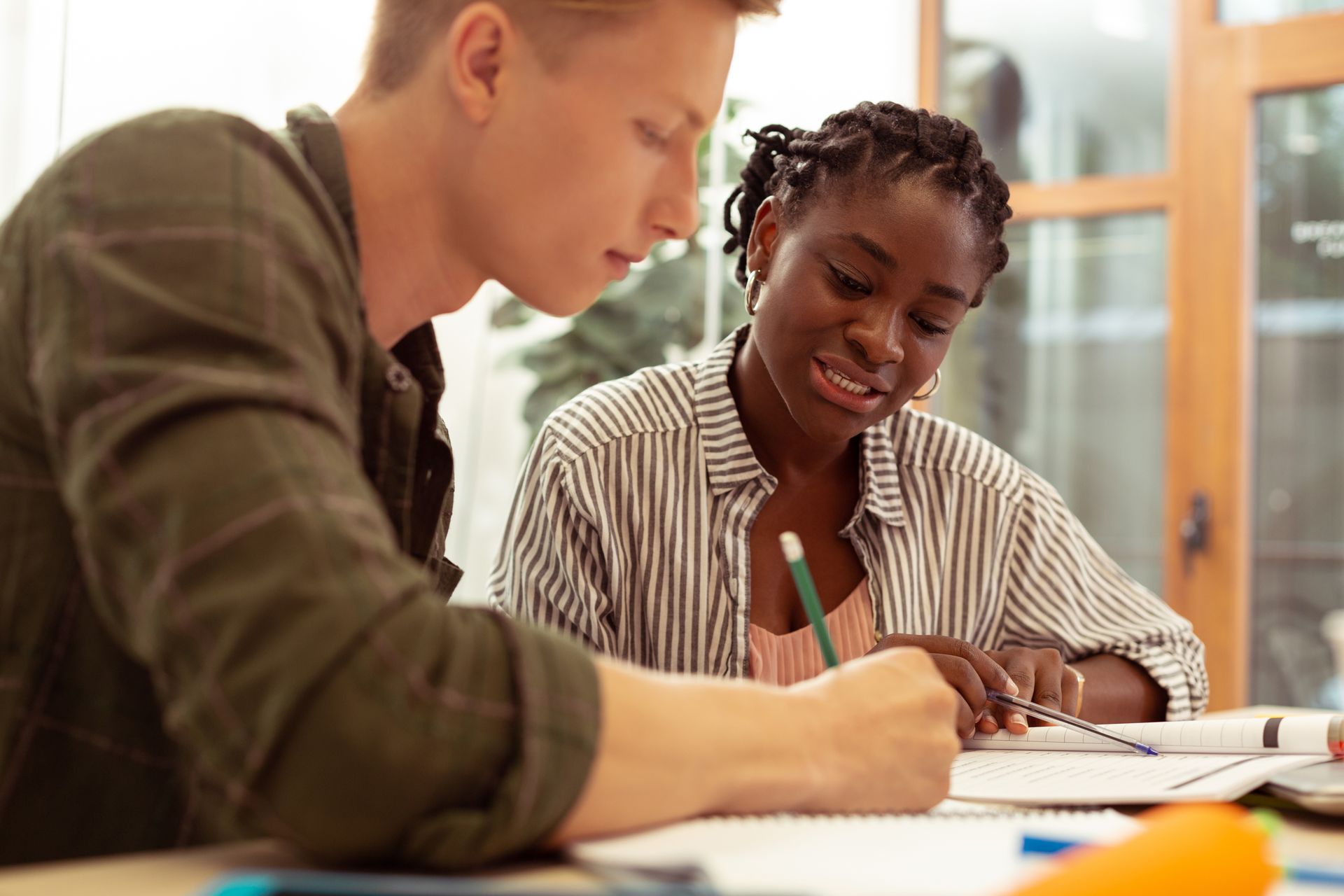Two people working together at a desk, one with blonde hair, the other with dark hair, writing in notebooks.