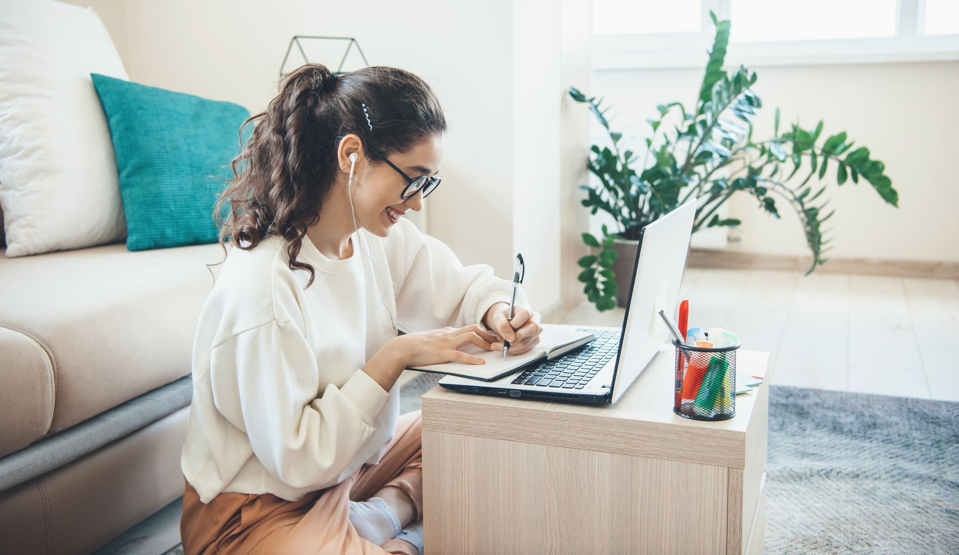 Woman writing in a notebook beside a laptop on the floor in a bright living room