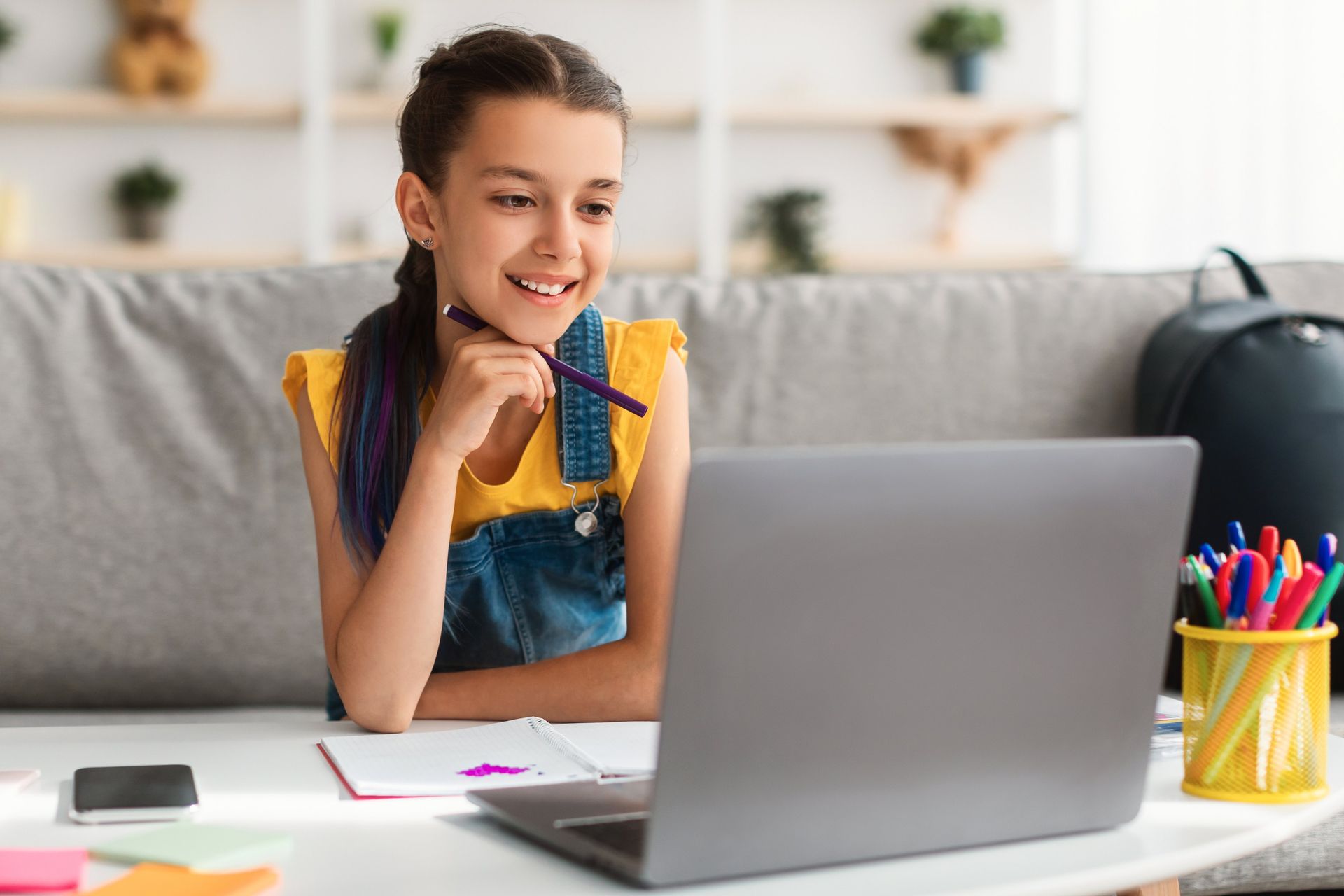 Smiling child on sofa using a laptop at a table with notebooks and pens nearby