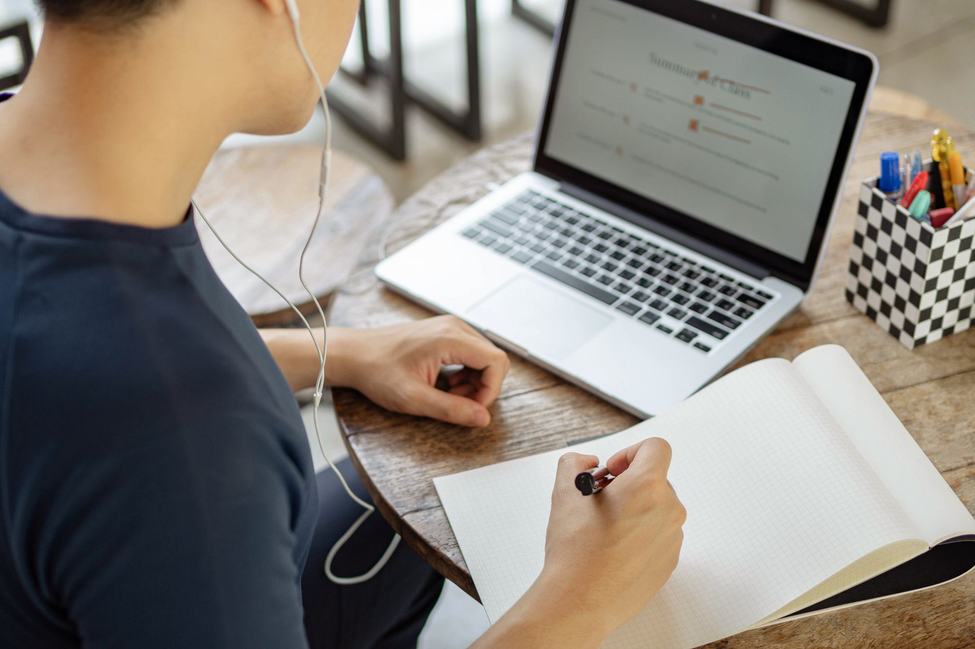 Person writing in a notebook beside a laptop on a desk, with earbuds in and a pen cup nearby.