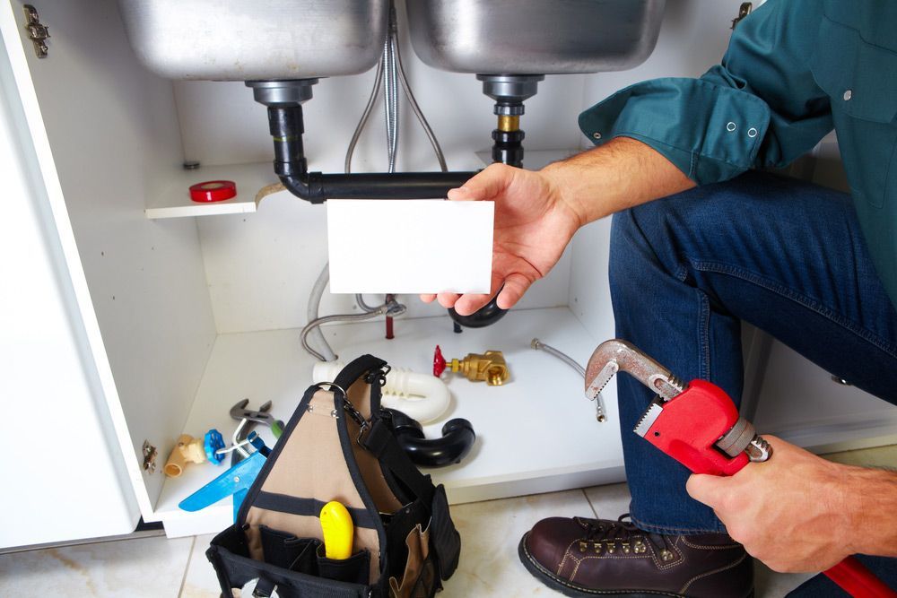 A Plumber Is Fixing A Sink In A Kitchen While Holding A Business Card — Pertzels Plumbing In Wagga Wagga, NSW