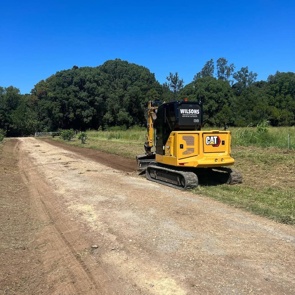 A Yellow Cat Excavator Is Driving Down A Dirt Road — Wilsons Excavations & Plant Hire in Tweed Heads, NSW