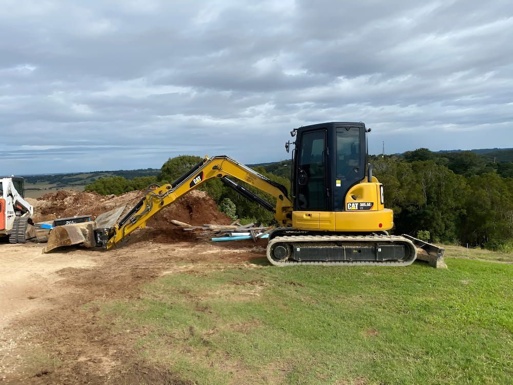 A Yellow And Black Excavator Is Sitting On Top Of A Grass Covered Field — Wilsons Excavations & Plant Hire in Myocum, NSW