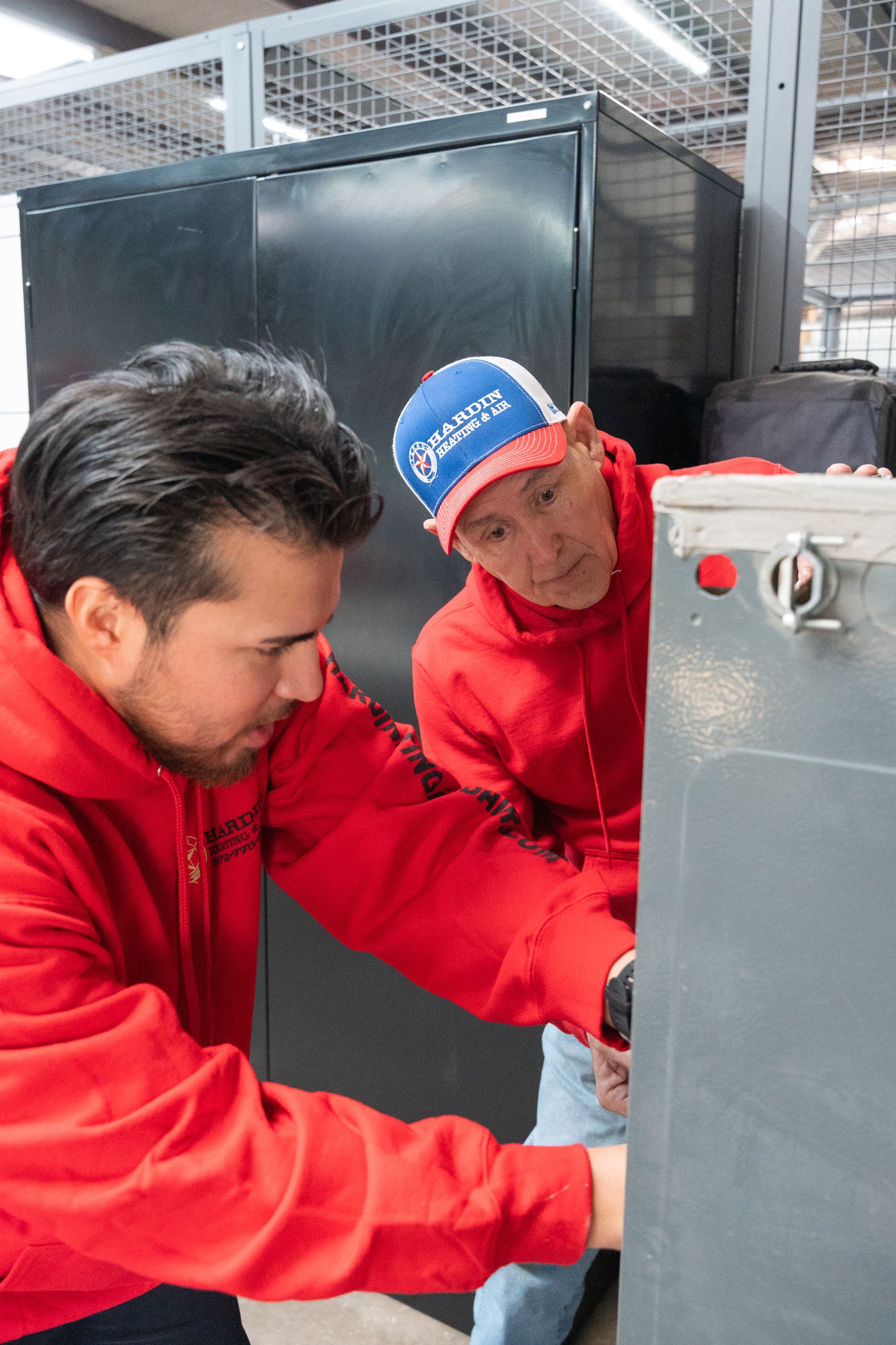 Two Hardin Heating & Air Technician in red hoodies working on a gray cabinet.