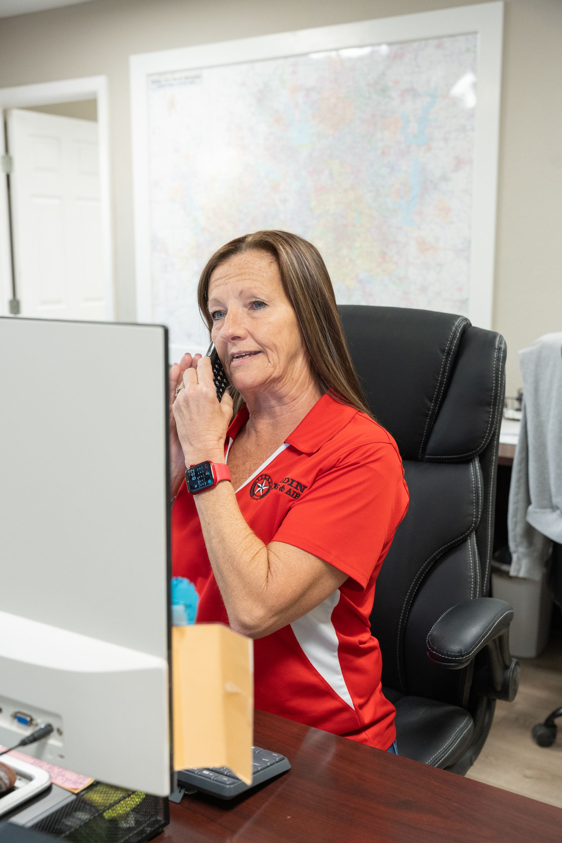 Woman in red shirt on phone at a desk, looking up, with a computer monitor and a map on the wall.