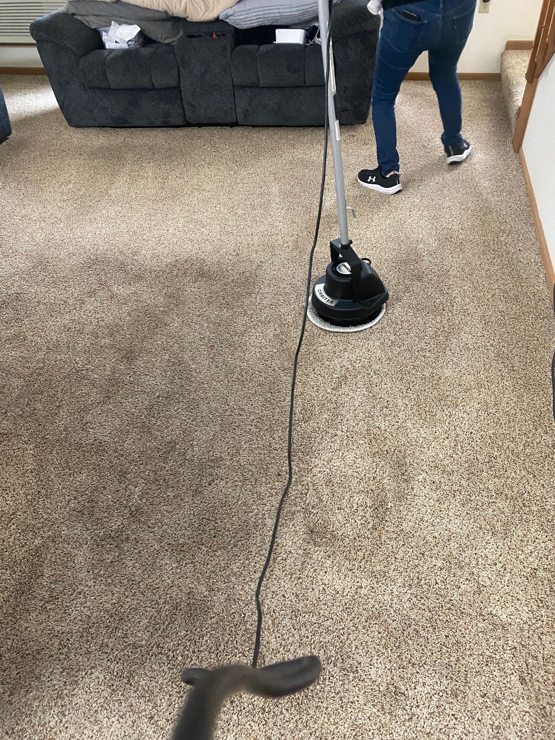 Person using a vacuum cleaner on a carpeted living room floor with a dark sofa nearby