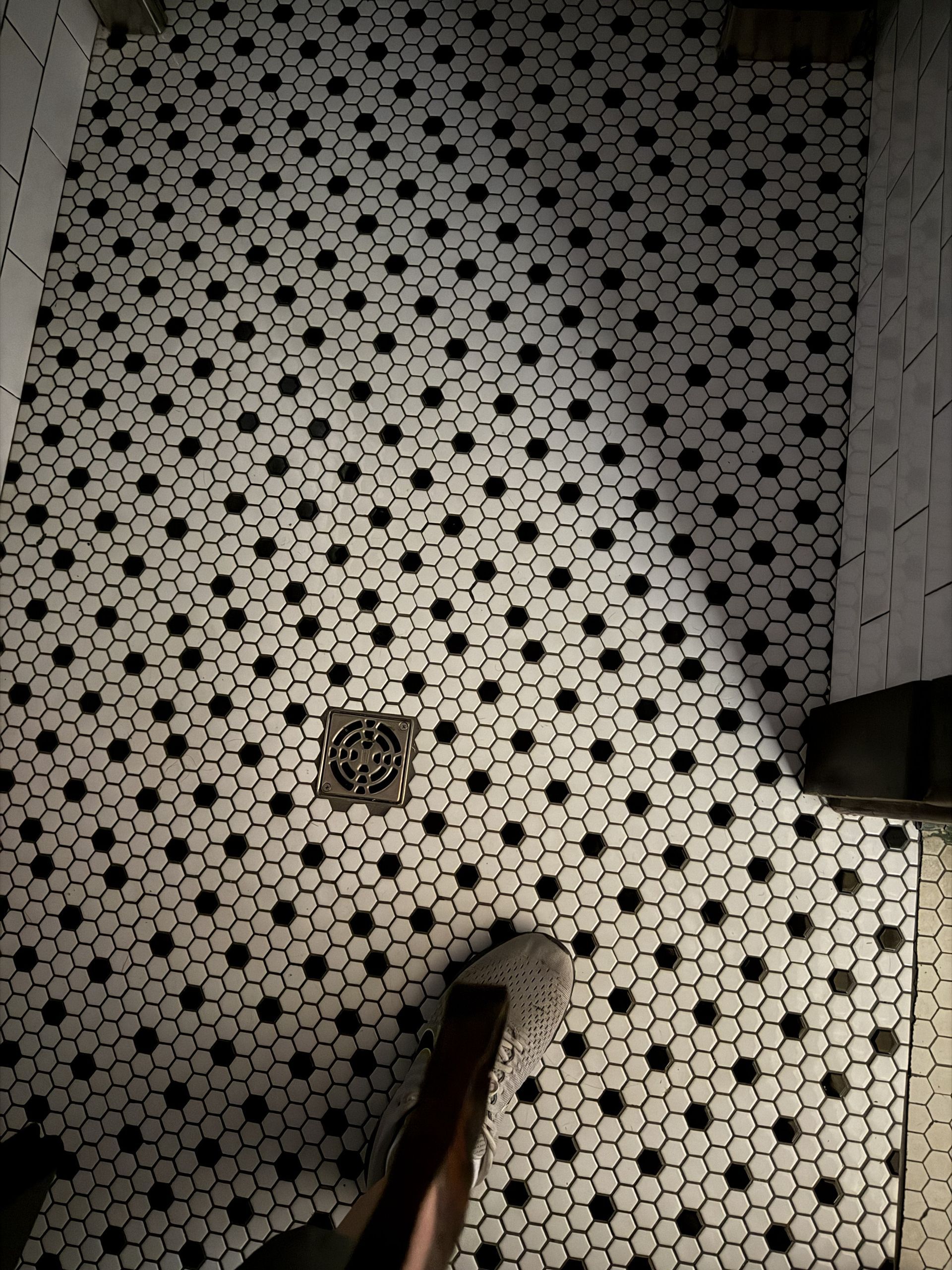 Black-and-white tiled floor with a drain, partially lit by a flashlight beam and a shoe at the bottom.