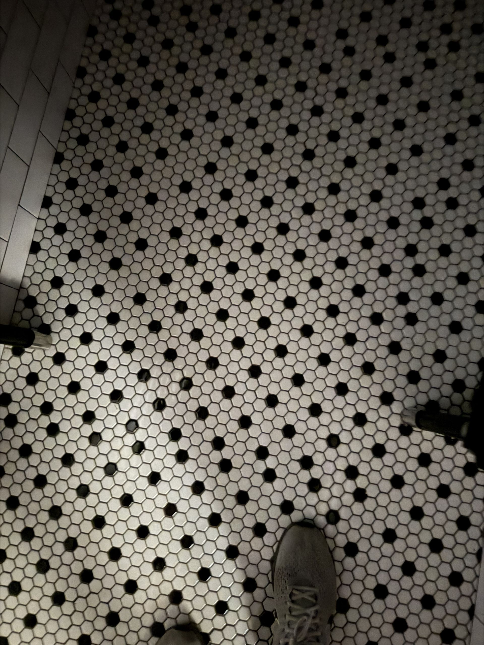 Top-down view of white hex tile floor with black dots and the tip of a sneaker at the bottom.