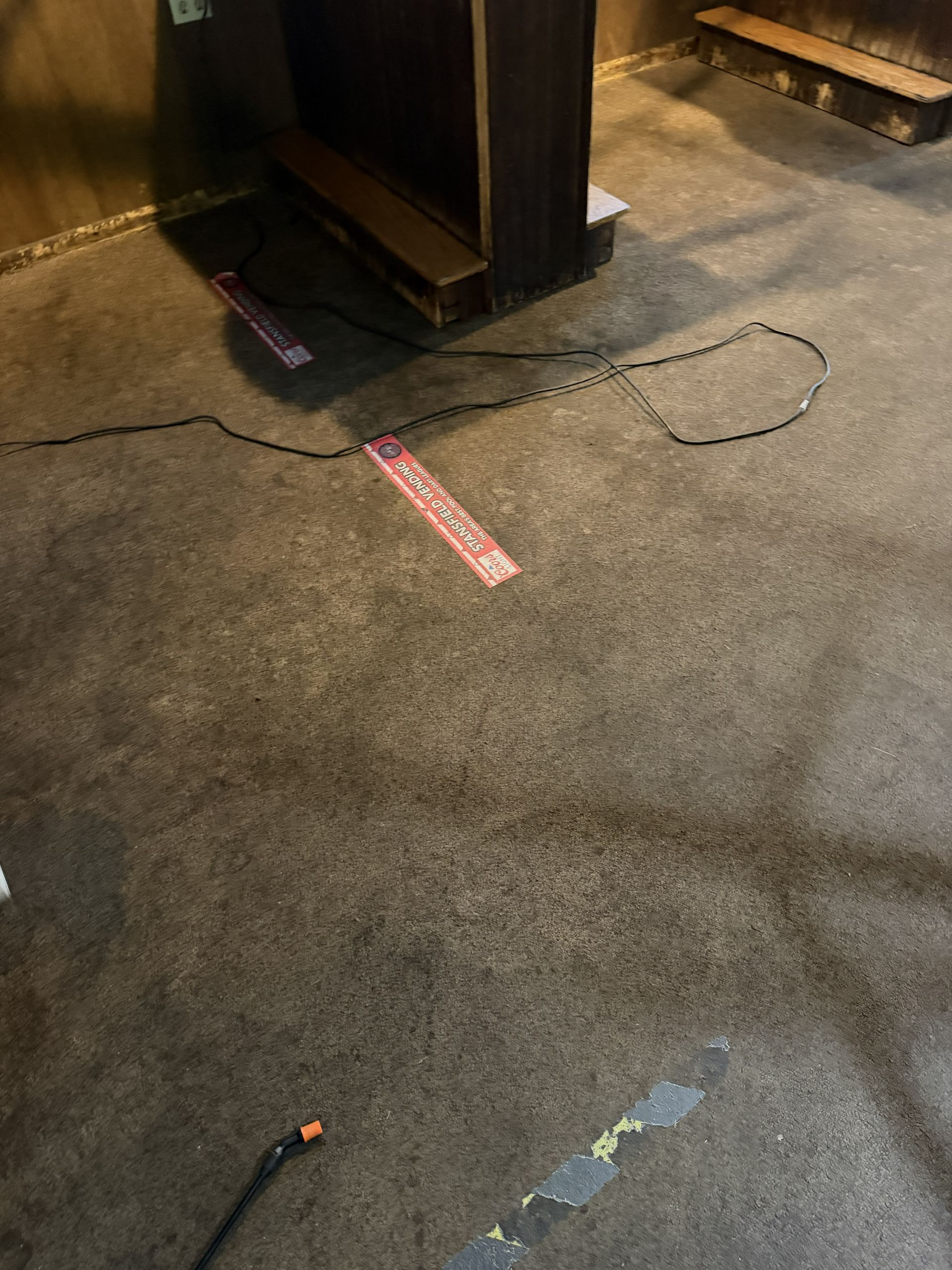 Carpeted floor with a small black cabinet, pink tape, and a cable lying across it.