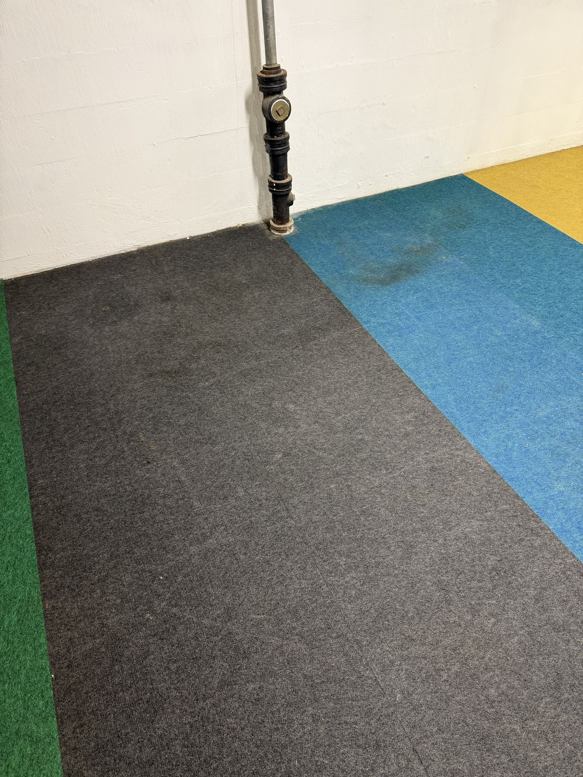 Colored carpet tiles meeting at a corner near a wall and pipe connection
