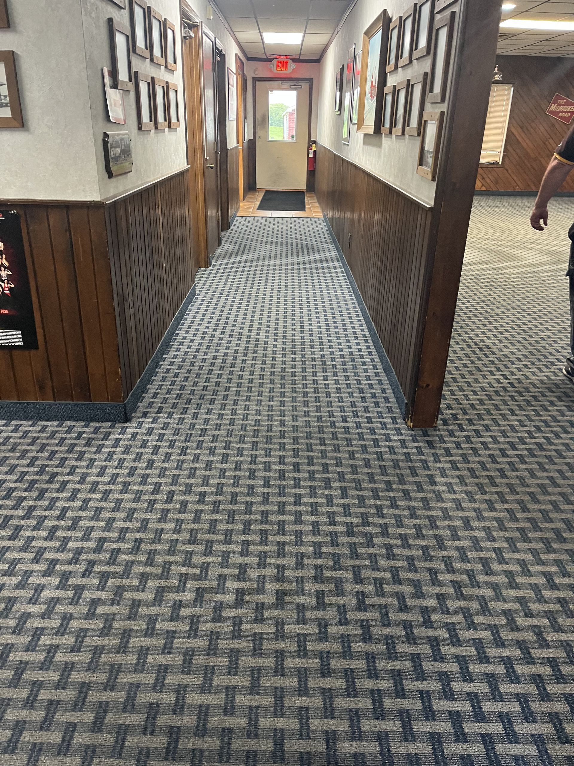 Carpeted hotel hallway with wood paneling and framed photos leading to a bright doorway