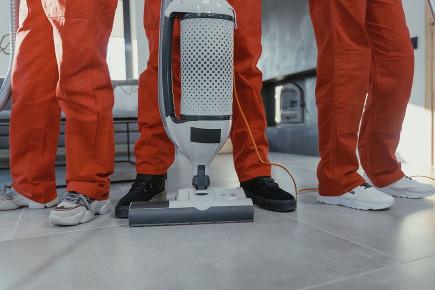 Three people in orange coveralls stand around a vacuum cleaner on a tiled floor.