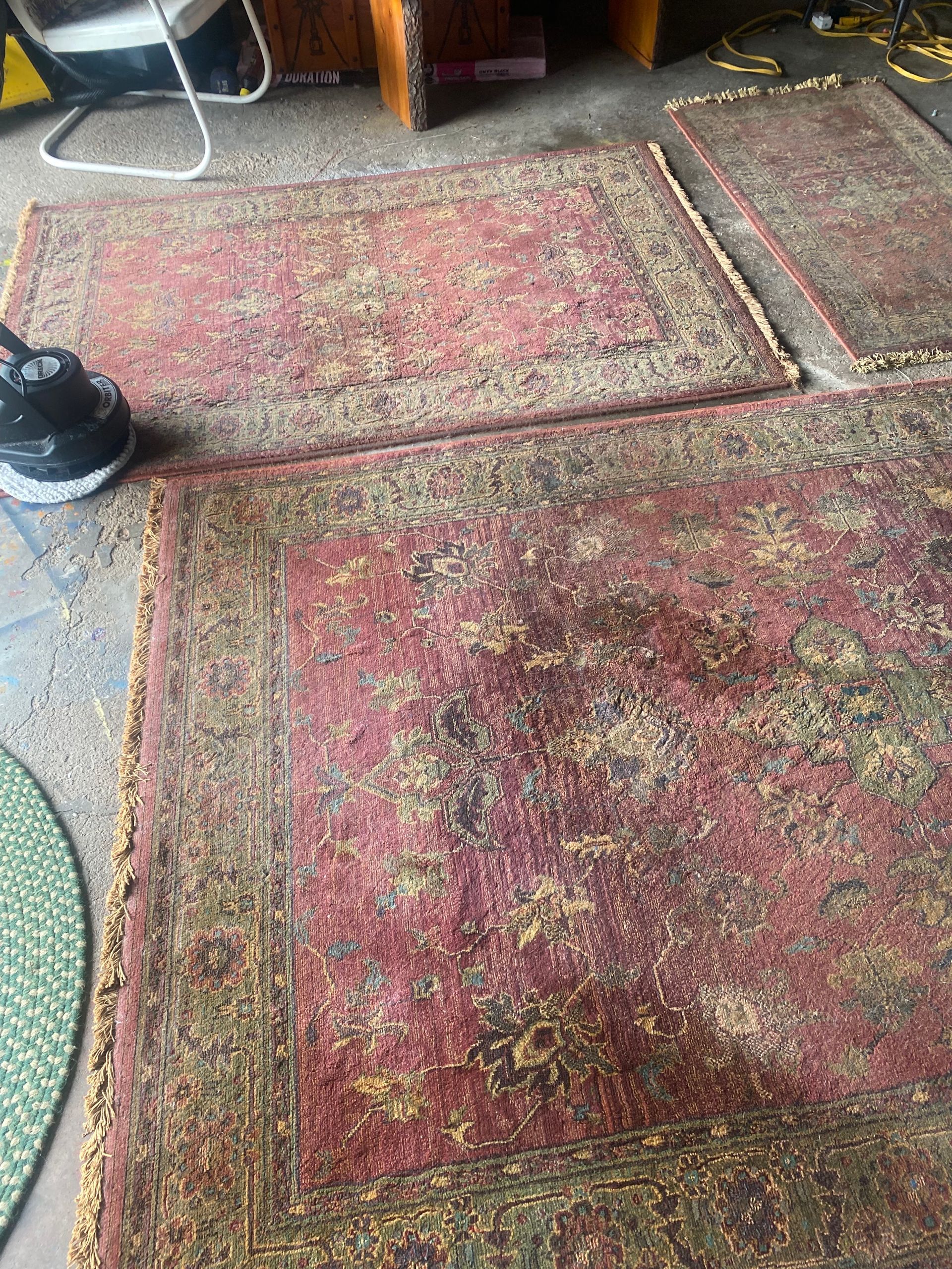 Worn red patterned area rug on a floor, with a black shoe at the left edge and furniture in the background