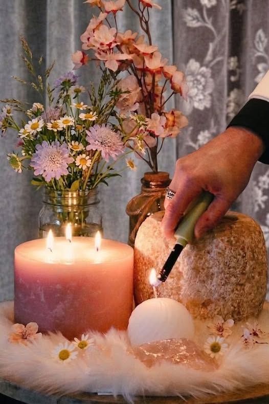 Een hand plaatst een witte kaars naast een brandende roze kaars, met bloemen en zachte decoraties op een warme tafel.
