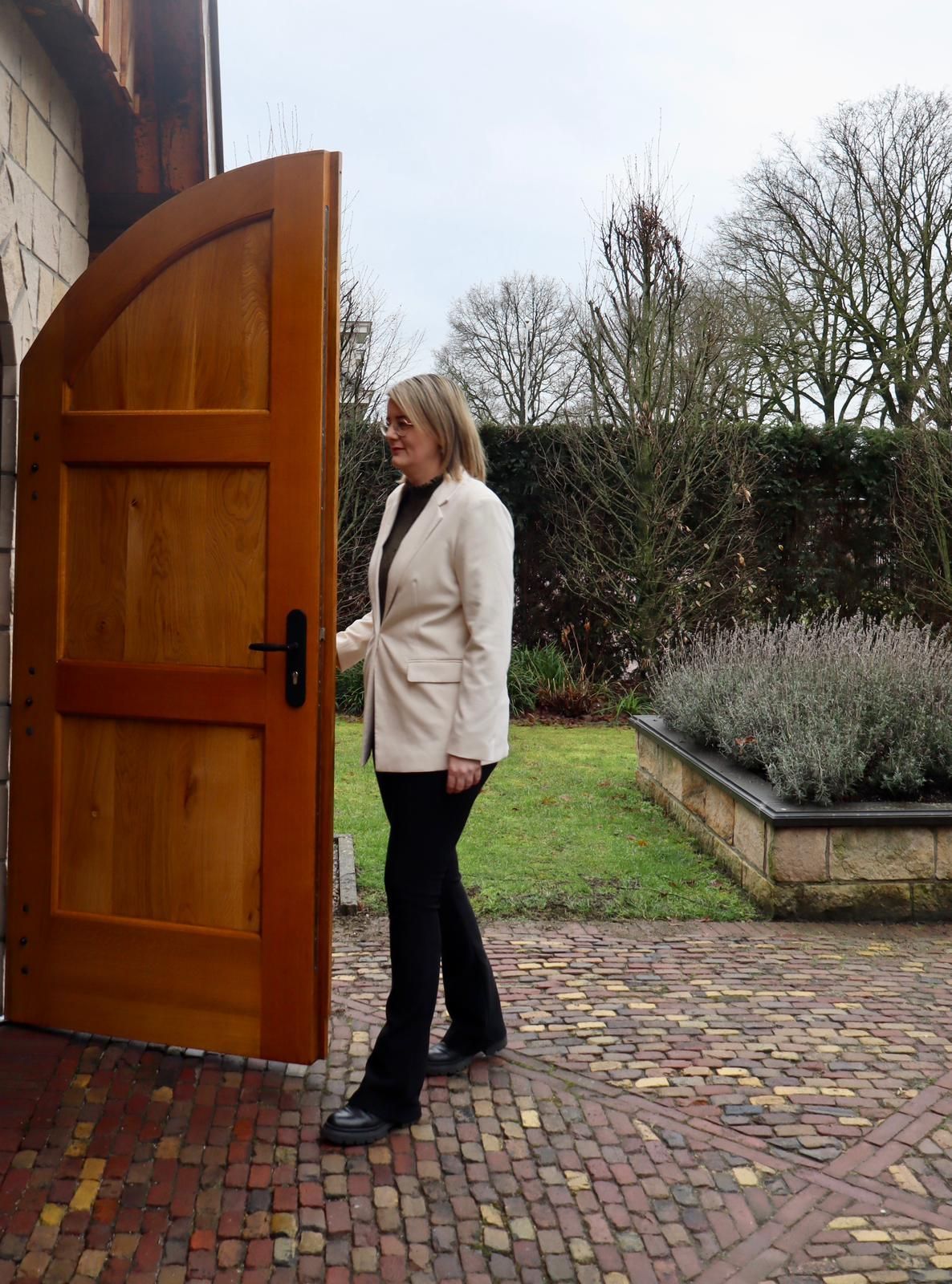 Een vrouw in een crèmekleurige jas opent een houten deur en loopt naar buiten, het stenen terras op.