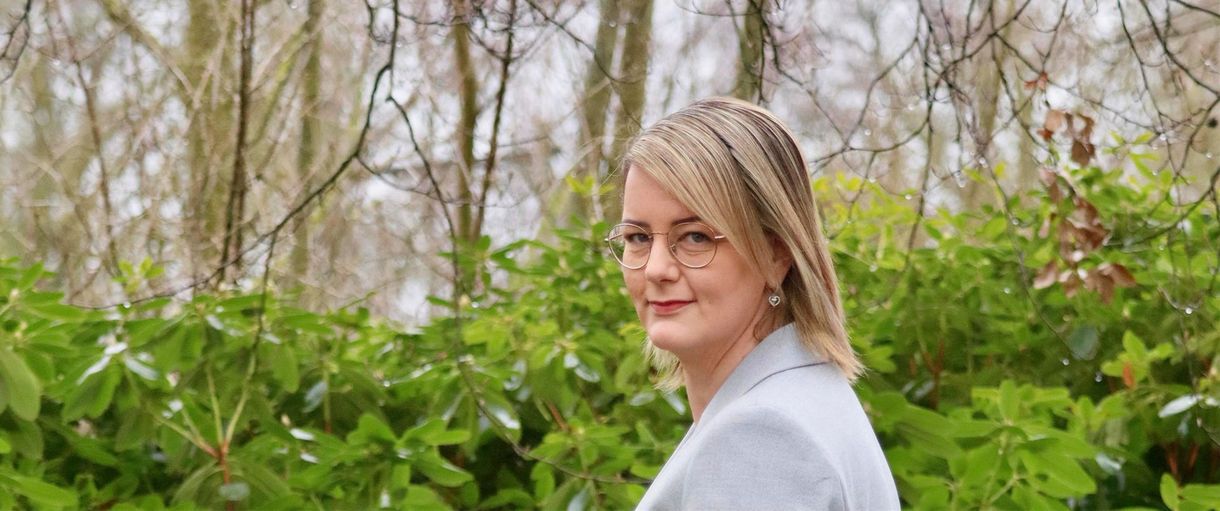 Vrouw met bril in een lichtgrijze blazer, lachend, voor groen gebladerte en bomen.