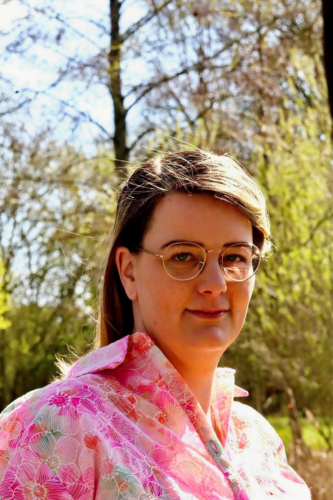 Een persoon met een bril en een roze bloemenblouse aan, buiten tussen de bomen, kijkt naar de camera.