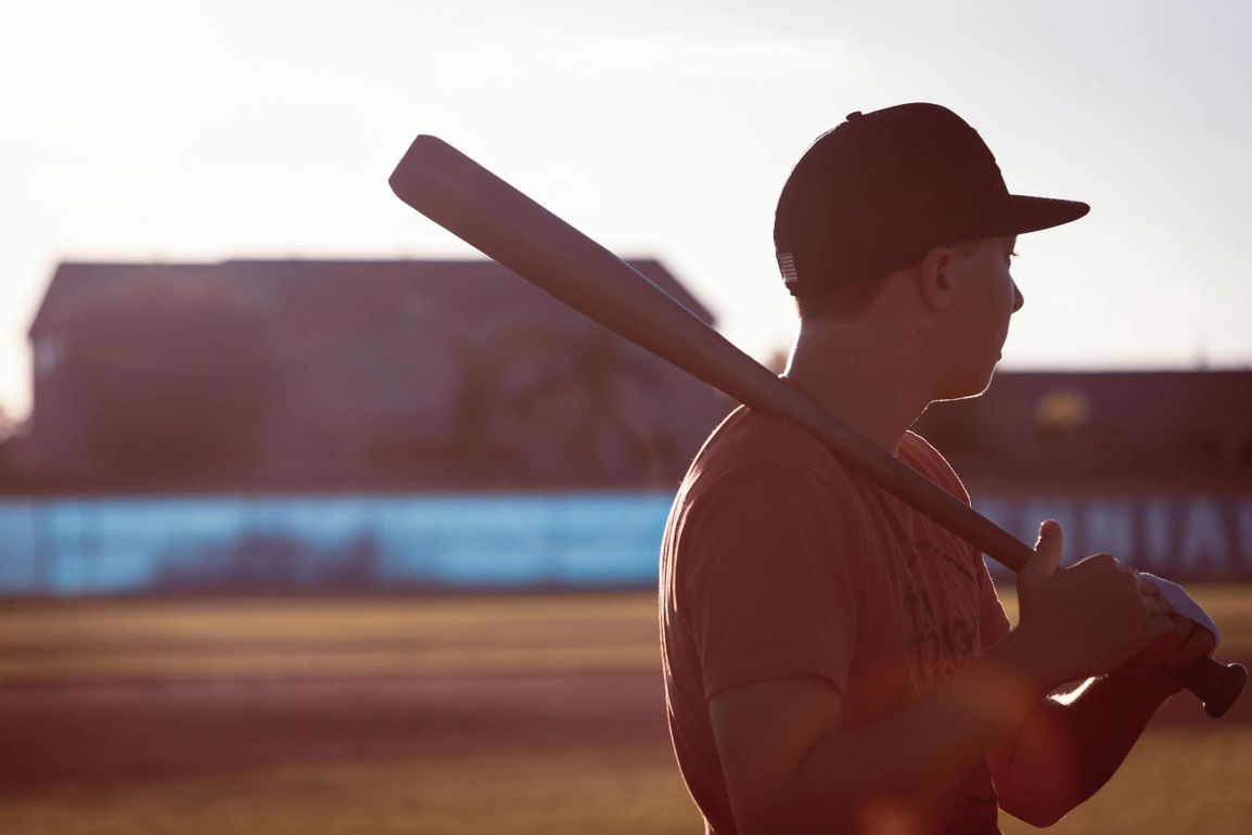 A person wearing a hat and orange shirt, holding a baseball bat on their shoulder on a baseball field.