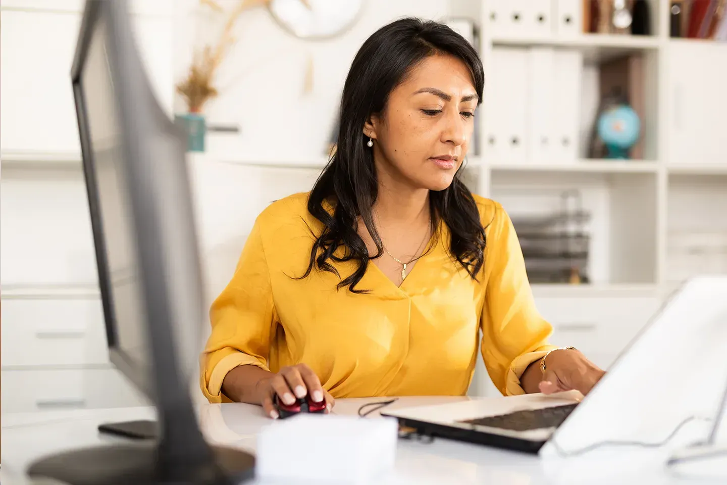 Business woman in a yellow blouse working at a desk with a laptop and desktop monitor