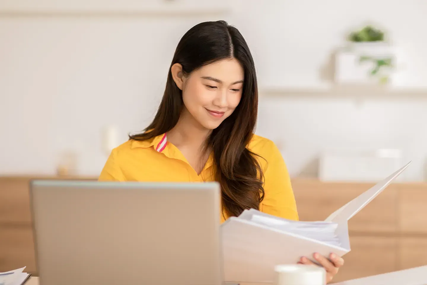 Beautiful Asian young woman using laptop computer and open document to reference her work.