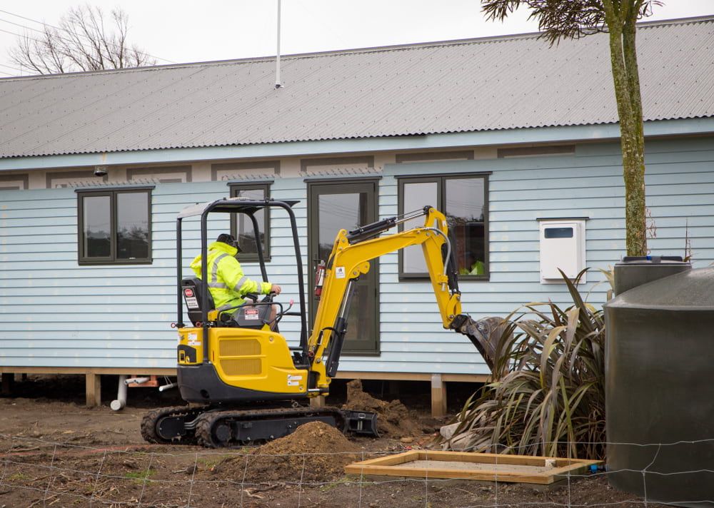 A Man is Driving a Small Excavator in Front of a House — Matt's Mini Machines in Bli Bli, QLD