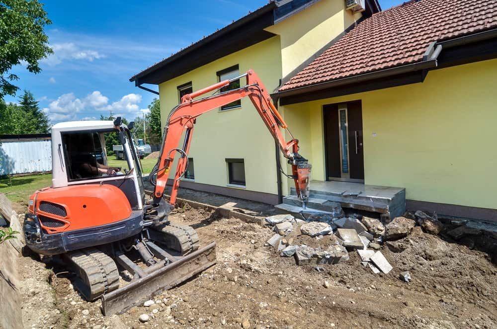 A Small Excavator is Sitting in Front of a Yellow House — Matt's Mini Machines in Buderim, QLD