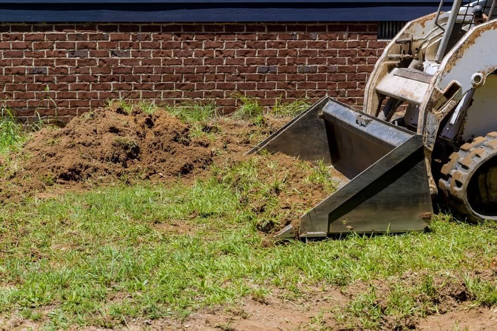 A Bulldozer is Digging a Hole in the Ground in Front of a Brick Building — Matt's Mini Machines in Palmwoods, QLD