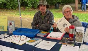 Two people at a table with papers and binders, one holding up a page outdoors under a canopy.