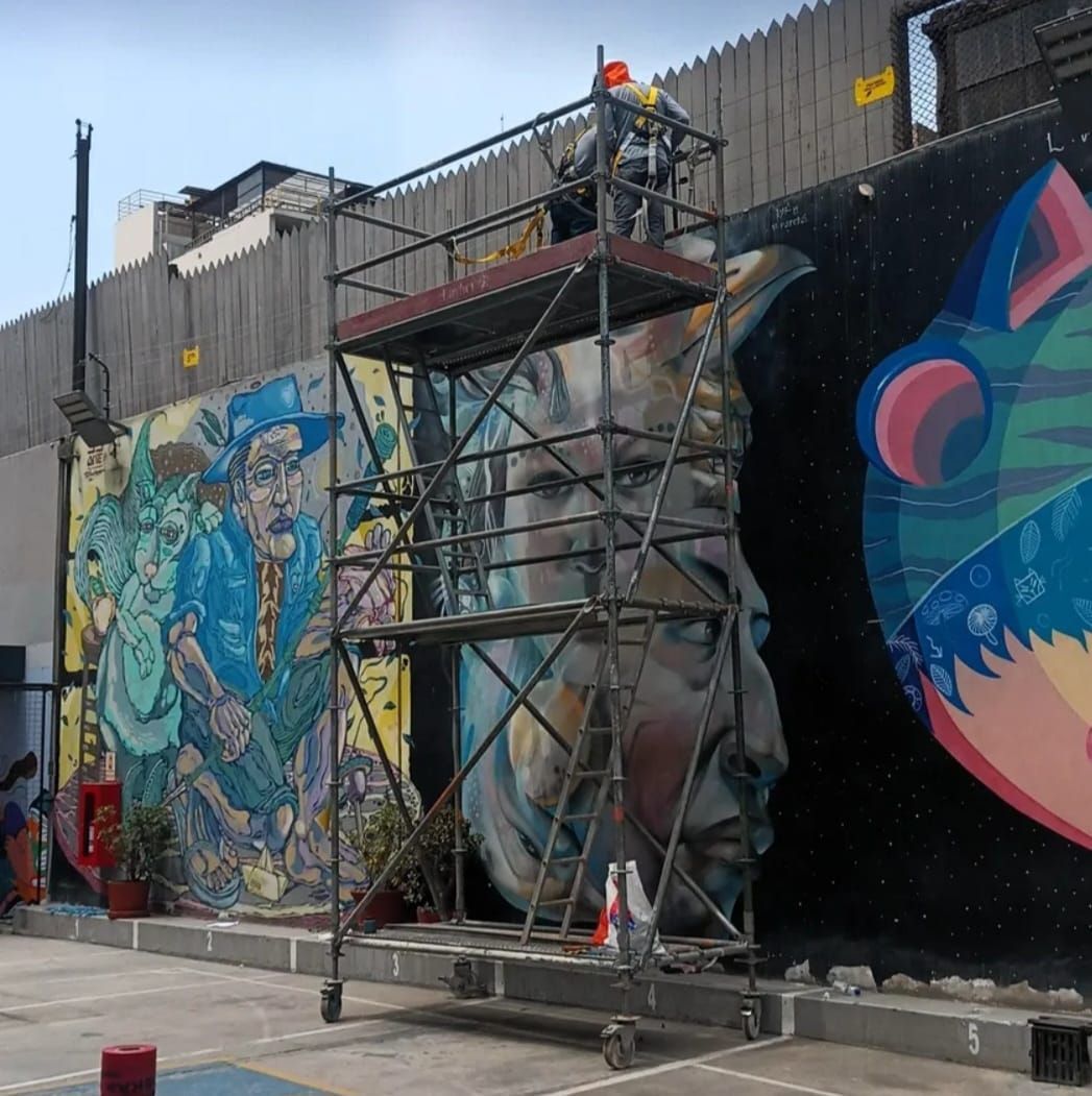 Un mural de grafiti está siendo reparado sobre andamios junto a una pared, con coloridos paneles de arte callejero.