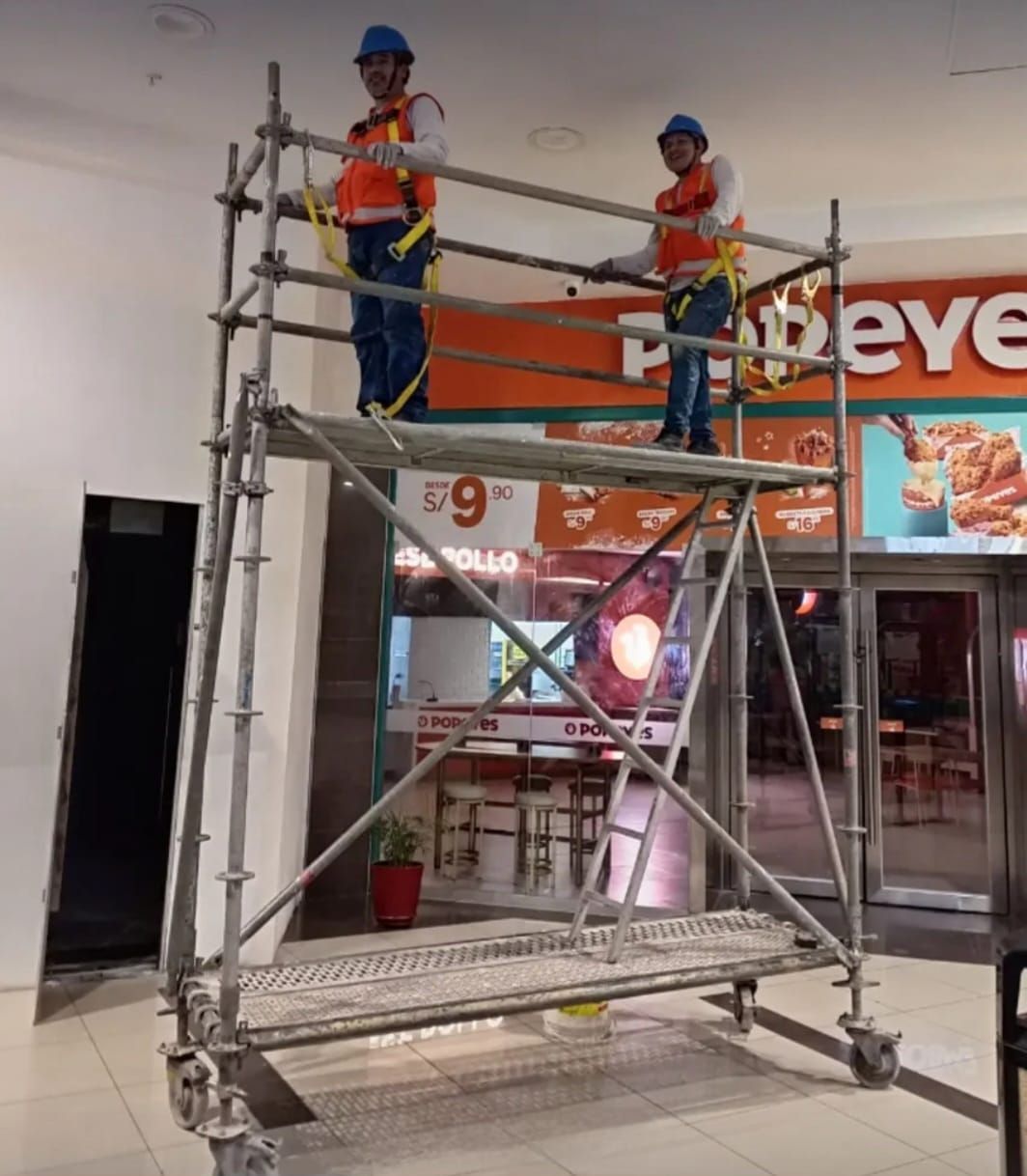 Dos trabajadores con cascos de seguridad en un andamio dentro de un centro comercial, cerca de la entrada de una tienda.