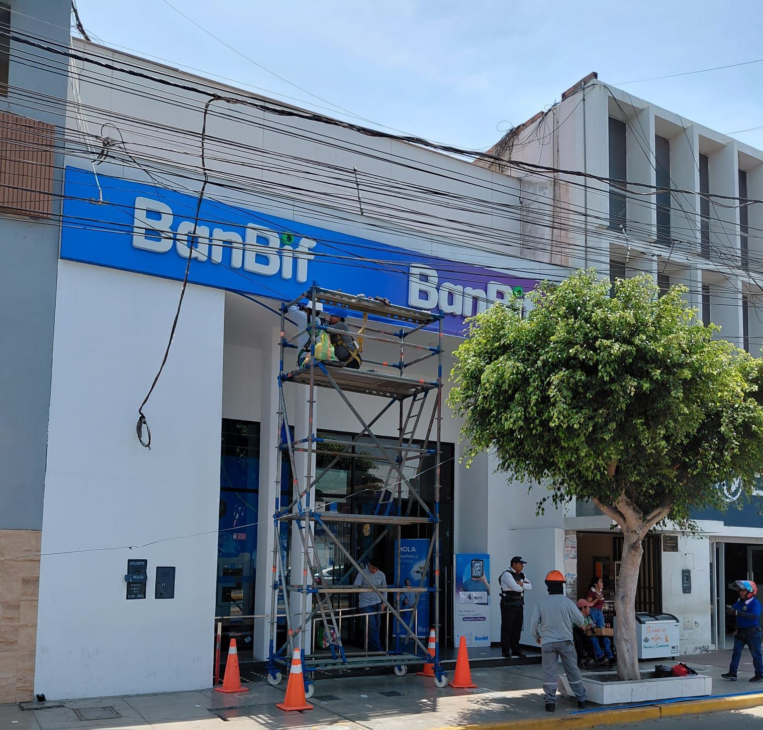 Fachada del edificio del banco con letrero azul, andamios y trabajadores junto a conos naranjas en una calle soleada.