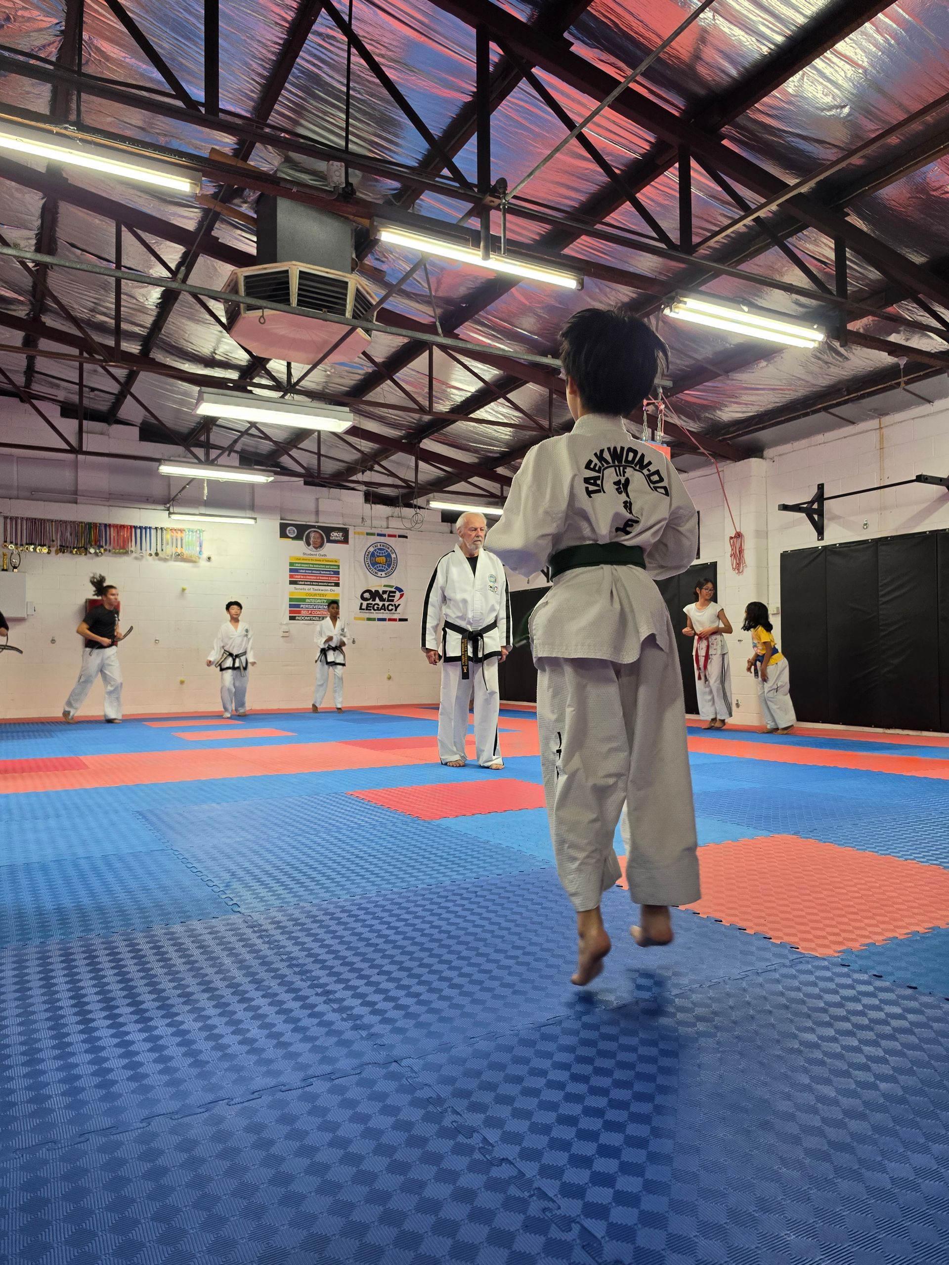 Children practicing martial arts on blue and red mats in a gymnasium with exposed ceiling beams.