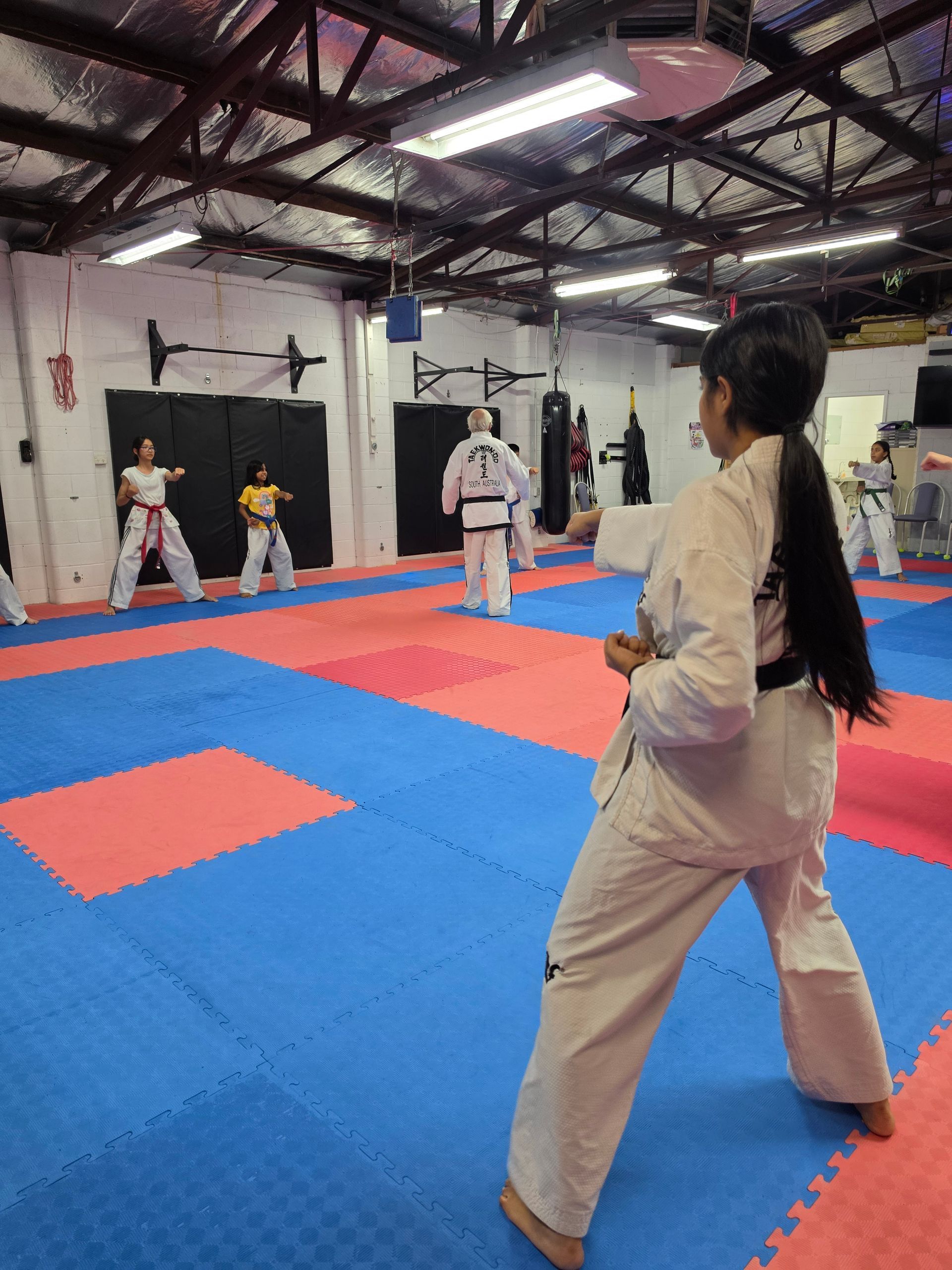 Martial arts class on red-and-blue mats in a gym, with students in white uniforms practicing drills