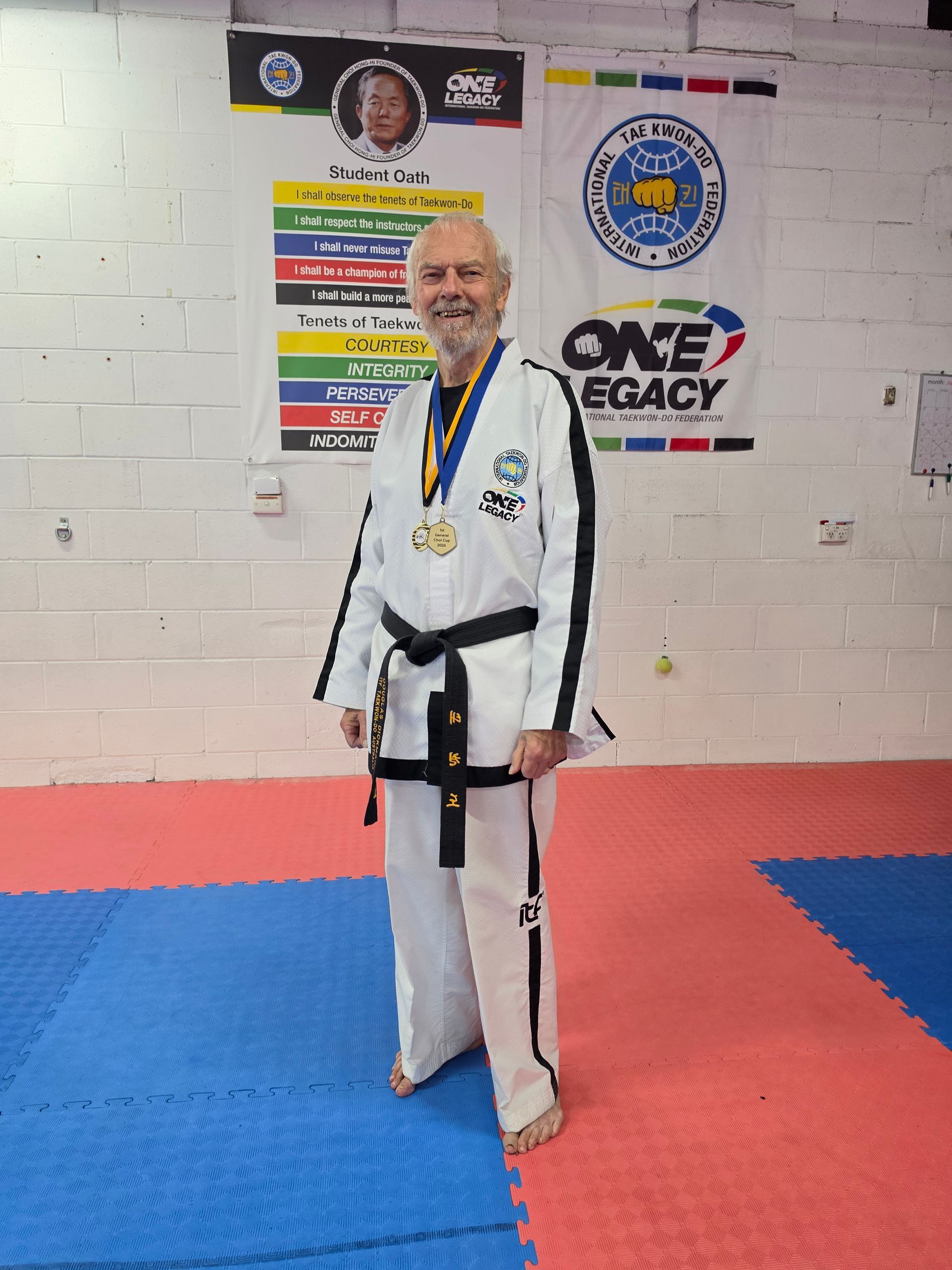 Martial artist in white gi with black belt standing on blue and red mats in a dojo, smiling at the camera